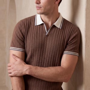 Puede incluir: Un polo marr&oacute;n acanalado con cuello blanco y ribete gris y blanco en el cuello y las mangas. La camisa tiene una cremallera corta y mangas cortas. El modelo est&aacute; apoyado en una pared beige.