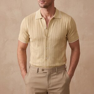 May include: A light tan polo shirt with short sleeves and a button-down front. The shirt has a textured pattern and white trim on the collar and sleeves. The model is also wearing tan pants.