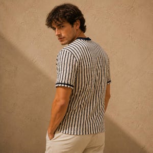 May include: A short-sleeved, striped shirt in cream and navy blue. The shirt features vertical navy stripes, a navy blue collar, and navy blue trim on the sleeves. The model is wearing khaki pants.