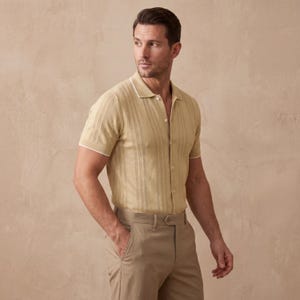 May include: A light tan short-sleeve polo shirt with a textured vertical stripe design and white trim. The shirt is buttoned and paired with tan trousers. The model is wearing a belt and has one hand in their pocket.