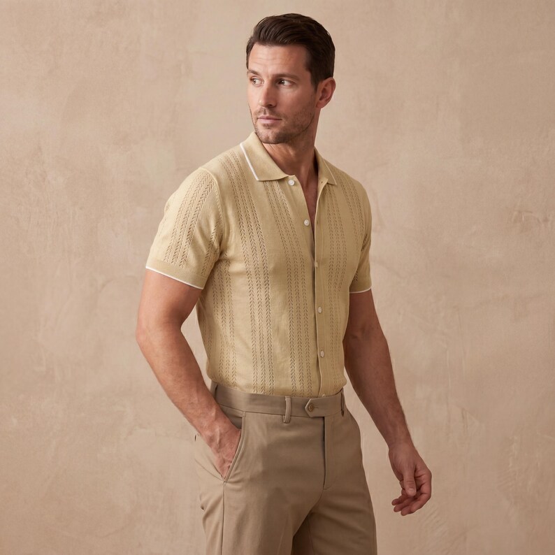 May include: A light tan short-sleeve polo shirt with a textured vertical stripe design and white trim. The shirt is buttoned and paired with tan trousers. The model is wearing a belt and has one hand in their pocket.