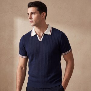 Puede incluir: Polo de manga corta azul marino con textura acanalada. La camisa tiene un cuello blanco con rayas rojas y blancas, y rayas a juego en los pu&ntilde;os de las mangas. La camisa tiene un dise&ntilde;o de cuello en V.