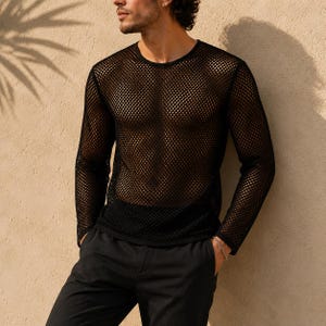 Op de afbeelding: Een zwart shirt met lange mouwen en een wijde, open gebreide mesh-constructie met een ronde halslijn. Het kledingstuk is semi-transparant en toont een ruitvormig rasterpatroon over de romp en mouwen, gecombineerd met een zwarte broek.