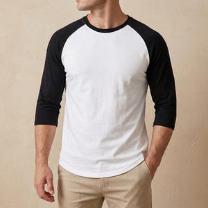 May include: A white baseball t-shirt with black sleeves. The shirt has a crew neck and three-quarter length sleeves. The model is wearing beige trousers.