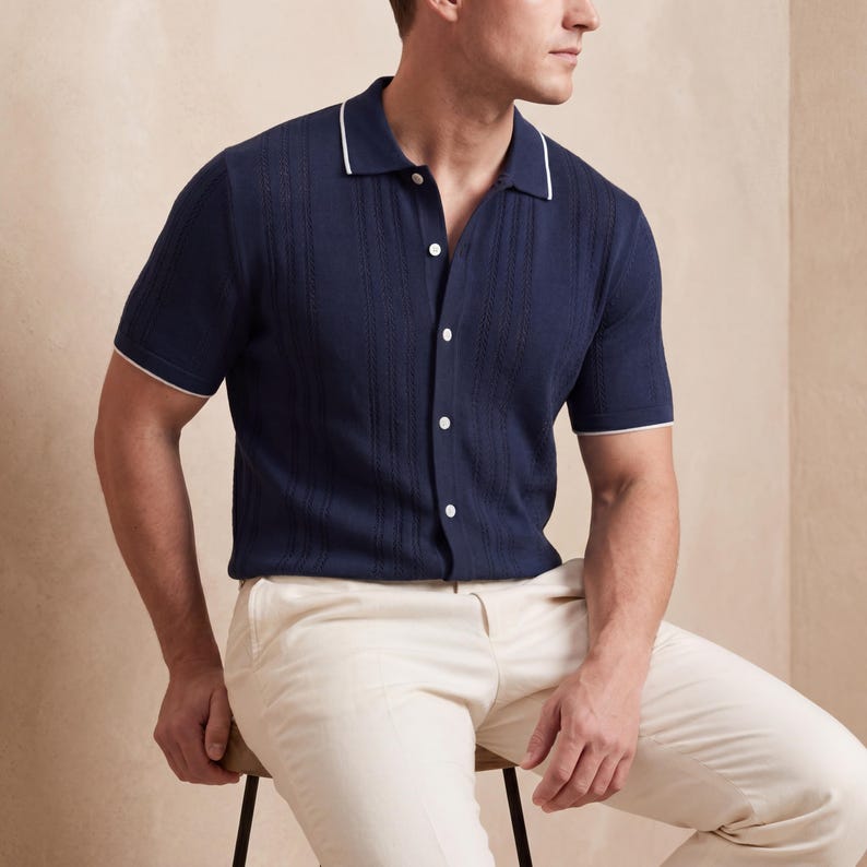 May include: Navy blue short-sleeve shirt with a ribbed texture, white trim on the collar and sleeves, and white buttons. The shirt is paired with cream-colored pants. The shirt is buttoned up.