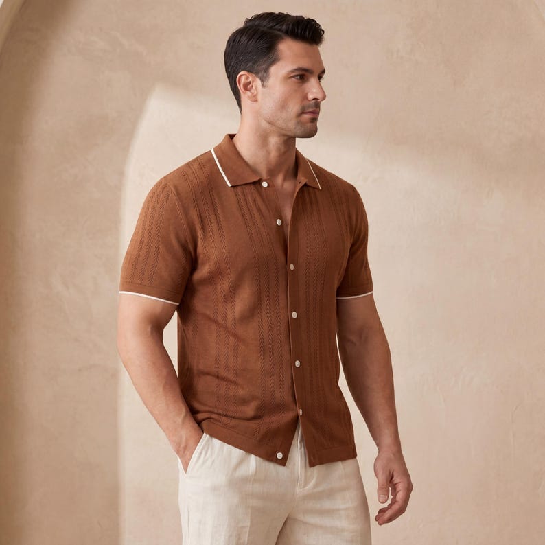 May include: A short-sleeved brown shirt with a textured knit pattern. The shirt features a white trim on the collar and sleeves, and is buttoned up. The model is wearing light-colored pants.