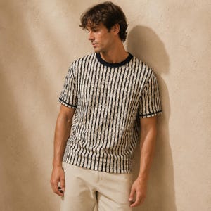 May include: A short-sleeved, striped t-shirt with a crew neck. The shirt features vertical black stripes against a cream-colored background. The neckline and sleeve cuffs are trimmed with a dark navy blue. The person is wearing beige pants.