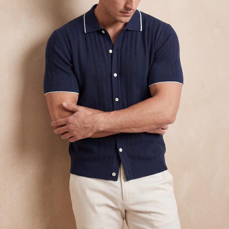 May include: Navy blue short-sleeve polo shirt with white trim on the collar and sleeves. The shirt has a button-down front and a textured design. The model is wearing cream-colored pants.
