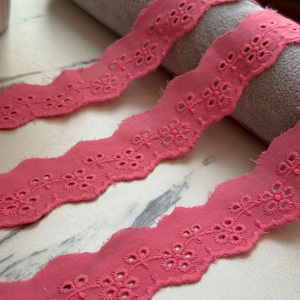 May include: A length of coral pink eyelet lace trim featuring a scalloped edge and a repeating floral embroidery pattern with small circular cutouts. The fabric strip is draped in three sections, showcasing the intricate needlework and texture.