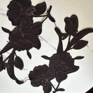 May include: Two black floral lace appliques featuring intricate embroidered petals, leaves, and stems. The designs showcase detailed stitching with textured centers, creating a symmetrical botanical pattern suitable for garment embellishment.
