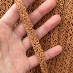 May include: A length of copper-colored crochet lace trim featuring a scalloped edge and a repeating openwork pattern. The trim is displayed against a matching copper-colored crochet fabric background, showcasing the intricate needlework and texture.