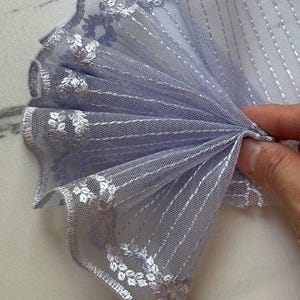 Puede incluir: Un borde de tela de malla color lavanda claro con un borde festoneado y un intrincado bordado plateado. El material transparente muestra un patrón floral repetitivo a lo largo del borde y líneas verticales cosidas a lo largo del textil.