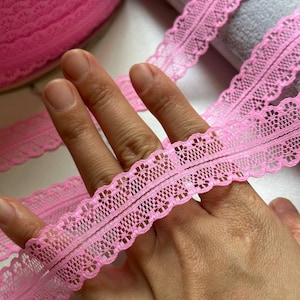 Puede incluir: Un carrete de cinta de encaje rosa brillante con un delicado patrón floral y bordes festoneados. El encaje está tejido con una textura de malla fina y se muestra como una tira continua, destacando el intrincado diseño calado y el color.