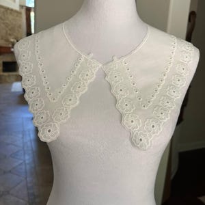 May include: A white detachable collar made of sheer fabric featuring intricate eyelet embroidery along the edges. The collar has a scalloped border with a repeating floral pattern and a series of small circular cutouts, displayed on a mannequin.