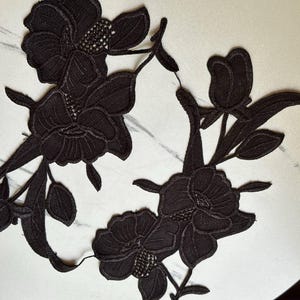 May include: Two black lace floral appliques featuring intricate embroidery of blooming flowers, buds, and leaves. The design showcases detailed stitching patterns on the petals and foliage, creating a textured, decorative motif for textile projects.