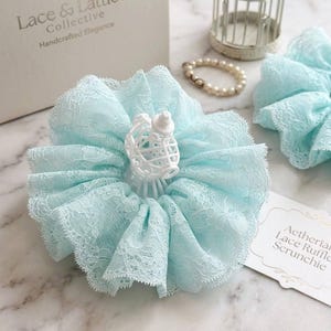 Op de afbeelding: Een lichtblauwe kanten scrunchie met een wit decoratief element. De scrunchie heeft een gerimpeld ontwerp van delicate kant. De afbeelding toont ook een bijpassende scrunchie, een parelarmband en een decoratieve vogelkooi.