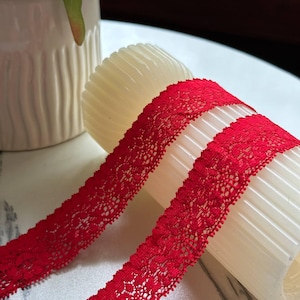 May include: A vibrant red lace trim ribbon featuring a delicate floral pattern with scalloped edges. The narrow fabric strip is wrapped around a cylindrical white spool, showcasing the intricate openwork texture of the red textile material.