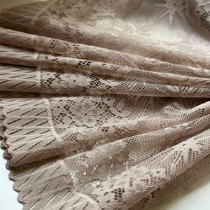 May include: A beige lace fabric featuring a detailed floral pattern and a scalloped edge. The material has a semi-sheer quality with a delicate, intricate weave throughout, showcasing a consistent texture and a soft, neutral color palette.