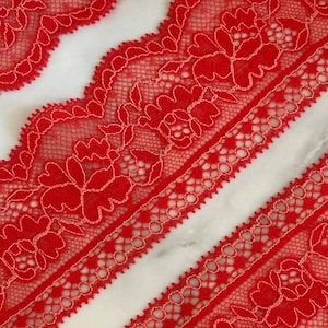 May include: A vibrant red lace trim featuring a repeating floral pattern with scalloped edges. The intricate design consists of delicate mesh netting and solid embroidered flower motifs, creating a continuous decorative border for sewing projects.