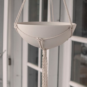 May include: Cream-coloured hanging planter with a rounded bowl shape, suspended by woven rope. The macrame design features a knotted pattern around the bowl and a long tassel. Suitable for displaying plants.