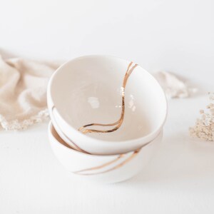 May include: Two stacked white ceramic bowls with a smooth, glossy finish. Each bowl features a distinct, hand-painted metallic gold line that curves across the interior and exterior surfaces, creating a continuous flowing design across the pieces.