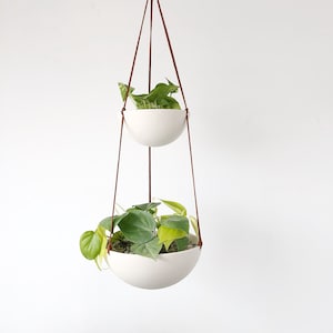 May include: Two white ceramic hanging planters with green plants, suspended from brown leather cords.
