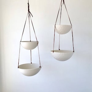 May include: Two white ceramic hanging planters with brown leather straps. The planters are suspended from the ceiling and are empty.