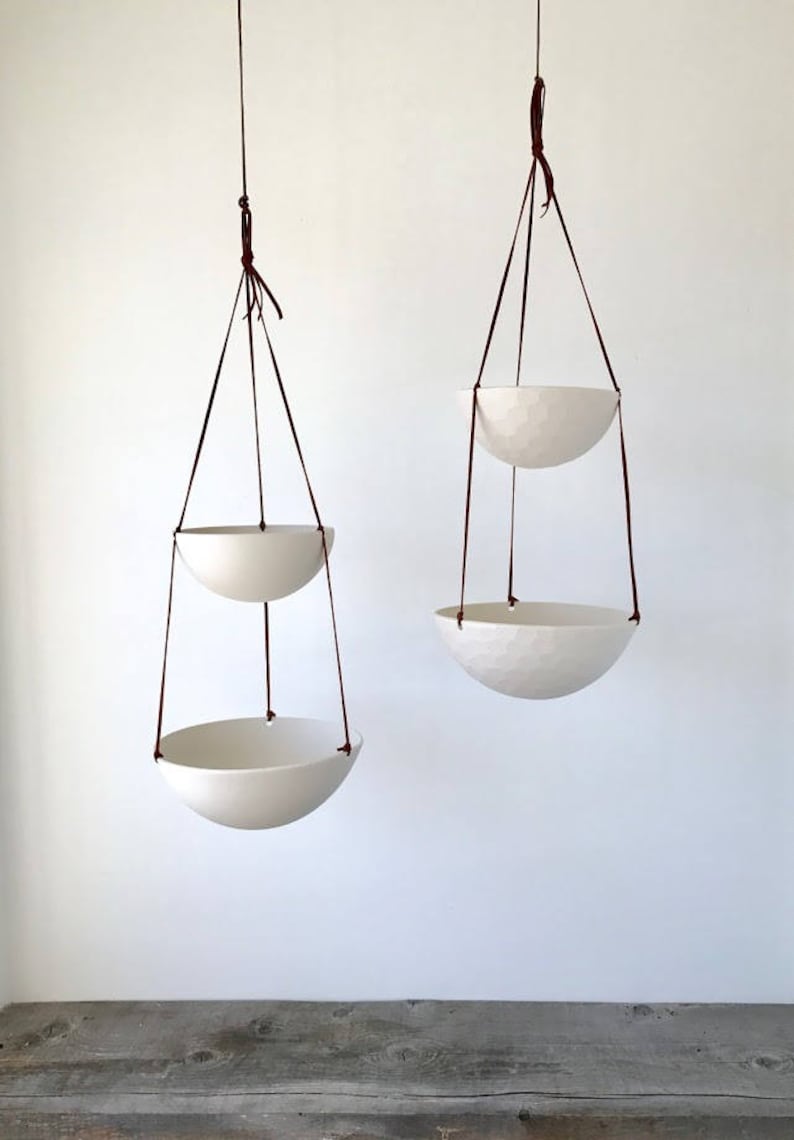 May include: Two white ceramic hanging planters with brown leather straps. The planters are suspended from the ceiling and are empty.