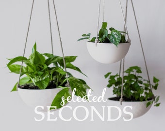 Op de afbeelding: Drie witte hangende plantenbakken met groene planten. De plantenbakken hangen aan touwen. De afbeelding bevat de tekst "selected SECONDS" in een sierlijk lettertype.