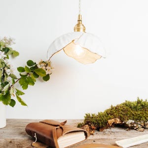 May include: A white, flower-shaped pendant light with a brass fixture hangs above a wooden table. A brown leather journal, a wooden plate, and a piece of moss are also on the table. The light is on, illuminating the space.