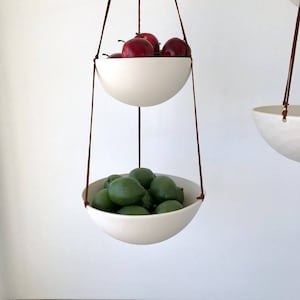 May include: Two white ceramic hanging bowls with brown leather cords. The top bowl is filled with red apples and the bottom bowl is filled with green limes.