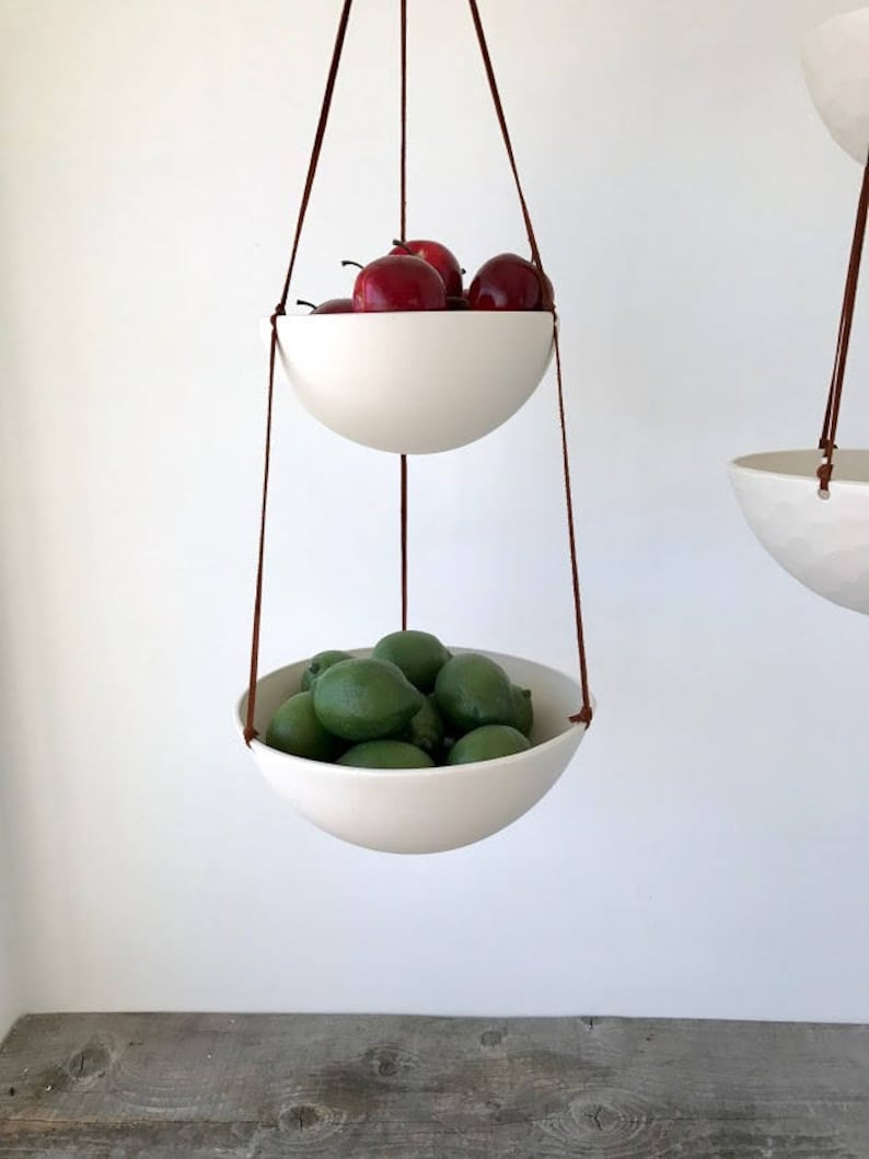 May include: Two white ceramic hanging bowls with brown leather cords. The top bowl is filled with red apples and the bottom bowl is filled with green limes.