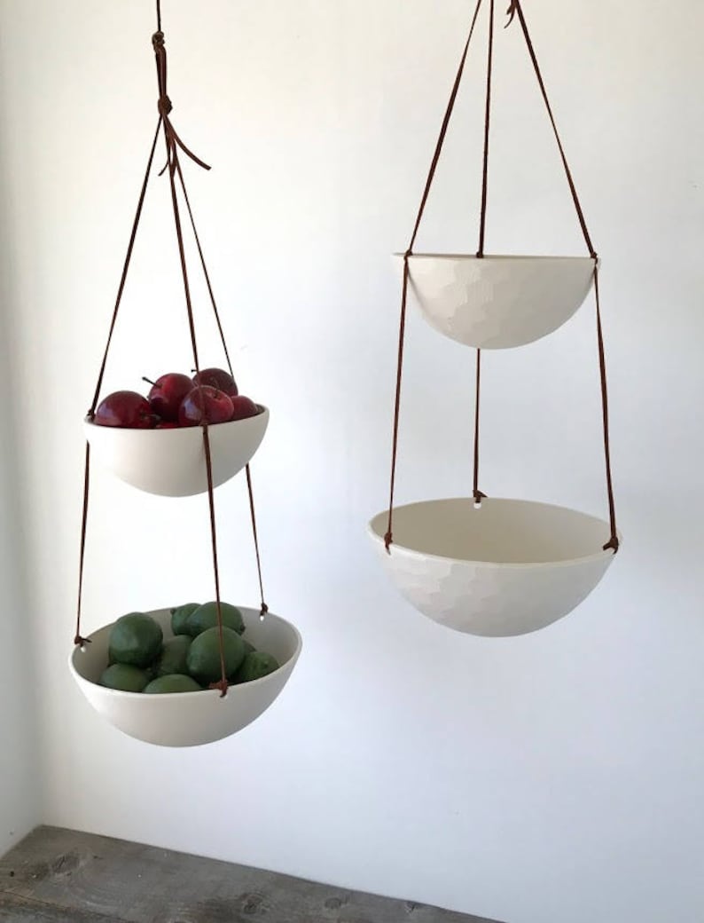 May include: Two white ceramic hanging bowls with brown leather straps. The top bowl is empty and the bottom bowl is filled with green limes.