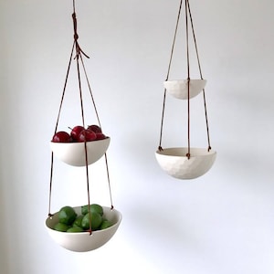 May include: Two white ceramic hanging bowls with brown leather straps. The top bowl is filled with red apples and the bottom bowl is filled with green limes.