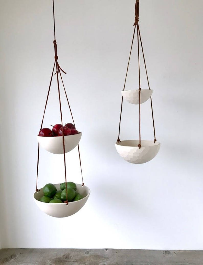 May include: Two white ceramic hanging bowls with brown leather straps. The top bowl is filled with red apples and the bottom bowl is filled with green limes.