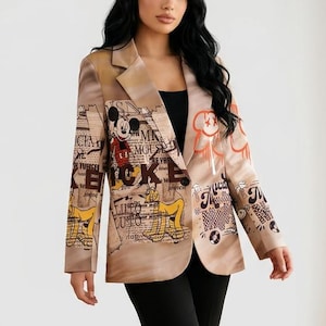 Puede incluir: Una blazer color beige con un estampado de Mickey Mouse y varios gráficos de texto. Se combina con pantalones negros y tacones negros. El estilo general es una mezcla de casual y artístico.