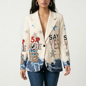 Puede incluir: Un blazer color hueso con un patrón de olas azules en el dobladillo, estrellas azules y plateadas dispersas, y texto gráfico que dice "SAY YES" y "52". El blazer incluye un pequeño gráfico de personaje animado y un botón negro.