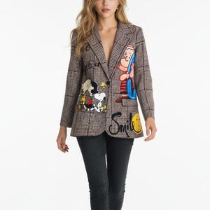 Könnte beinhalten: Ein brauner Blazer mit Karomuster, Cartoon-Charakter-Grafiken und dem Wort "Smile" in schwarzer Schrift. Der Blazer wird mit schwarzen Jeans und schwarzen Stiefeletten getragen. Der Blazer hat ein klassisches Revers.