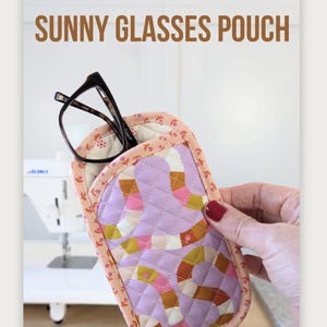 May include: A quilted fabric pouch for eyeglasses featuring a lavender background with a winding snake pattern in shades of pink, orange, and yellow. The pouch has a peach-colored floral trim and contains a pair of black-framed glasses. Text reads SUNNY GLASSES POUCH and MA TANTE QUILTING.