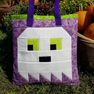 K&ouml;nnte beinhalten: Lilafarbene Tragetasche mit einem wei&szlig;en Geistergesicht auf der Vorderseite. Der Geist hat gr&uuml;ne Augen und einen schwarzen Mund. Die Tasche hat einen gr&uuml;nen und wei&szlig;en Candy-Corn-Besatz. Die Tasche liegt auf einem Bett aus gr&uuml;nem Gras. Der Text "Ghost Tote Bag" befindet sich oben im Bild. Der Text "www.sewfun.com #116" befindet sich unter dem Titel.