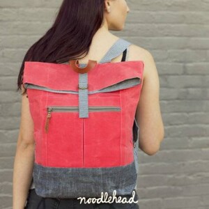 May include: A red foldover style backpack with a zippered front pocket and a black and white checkered strap. The backpack is made of canvas and has a leather accent. The text "RANGE BACKPACK" and "a foldover style backpack with zippered front pocket" is at the top of the image. The text "noodlehead" is at the bottom of the image.