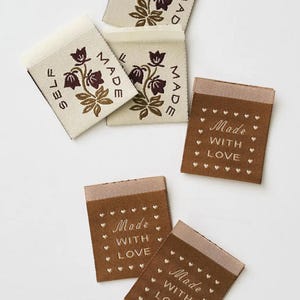 May include: A collection of rectangular fabric clothing labels. Three off-white labels feature a dark brown floral design and the text SELF MADE. Two brown labels feature the text MADE WITH LOVE surrounded by a border of small white hearts.