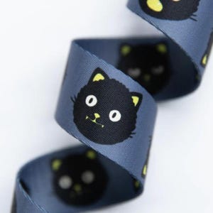 May include: A blue ribbon featuring a repeating pattern of black cat faces with white eyes and yellow inner ears. The cat faces are stylized with simple circular eyes and small yellow accents on the ears, arranged in a continuous design on the fabric.