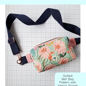 May include: A quilted Alpen belt bag with a floral pattern in pink, green, and peach. The bag has a navy blue adjustable strap with gold-colored hardware. The text "Alpen Belt Bag" is at the top of the image. The bag is a pattern with an interior pocket.