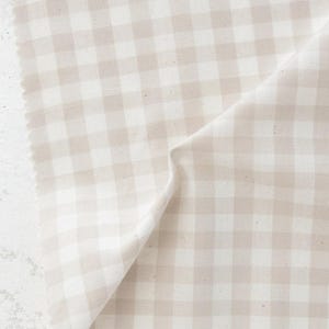 May include: A close-up of a beige and white gingham fabric. The fabric is folded, revealing the texture and pattern of the woven material. The edges of the fabric are visible, showing the raw, unfinished edge.