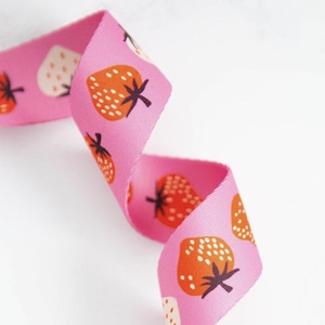 May include: Pink ribbon with a repeating pattern of orange and white strawberries. The ribbon is folded and twisted, showing the pattern from different angles.