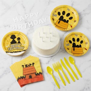 May include: A birthday party set with yellow plates, napkins, and cutlery featuring Snoopy and Woodstock designs. Includes a white iced cake with candles and a "Happy Birthday" banner.