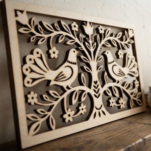 Könnte beinhalten: Ein rechteckiges Wandbild aus hellem Holz mit einem lasergeschnittenen Motiv von zwei Vögeln, die auf gegenüberliegenden Seiten eines zentralen Baumes sitzen. Das filigrane Ausschnittmuster zeigt stilisierte Blätter, kleine Blüten und Äste.