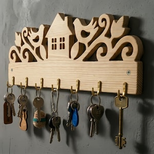 May include: Wooden key holder with a house and bird design. The light-colored wood has decorative cutouts and brass hooks for keys. Several keys hang from the hooks. A decorative and functional home decor item.
