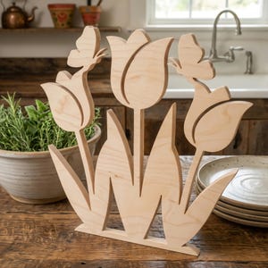 Könnte beinhalten: Holzdekoration mit Tulpen und Schmetterlingen. Die Naturholzskulptur zeigt drei Tulpen mit Schmetterlingsakzenten. Die Blätter und Stiele sind ausgeschnitten, wodurch ein offenes Design entsteht. Die Skulptur steht auf einem Holztisch.